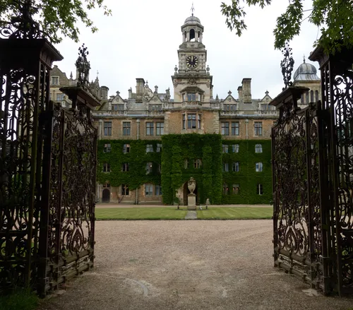 This architectural photograph captures the grand entrance gates and the impressive facade of Thoresby Hall, a historic country house located in Nottinghamshire, England, United Kingdom. Taken in the late afternoon during the summer, the image shows the ornate wrought iron gates framing a view of the ivy-clad Thoresby Hall, with its tall clock tower and elegant stonework standing out against the summer greenery. The carefully manicured lawn and the symmetrical design of the hall emphasize the splendour of this notable landmark, which is recognised as one of the prominent country houses in the region. The photograph highlights both the beauty of Thoresby Hall and the rich architectural heritage found in Nottinghamshire.